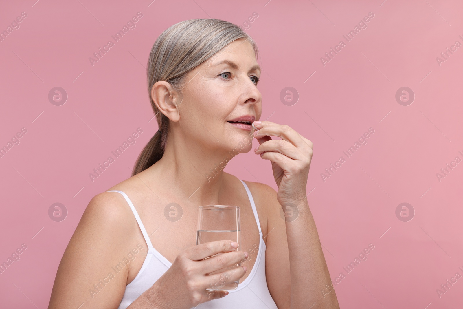 Photo of Beautiful woman with glass of water taking vitamin pill on pink background