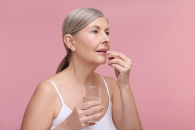 Beautiful woman with glass of water taking vitamin pill on pink background