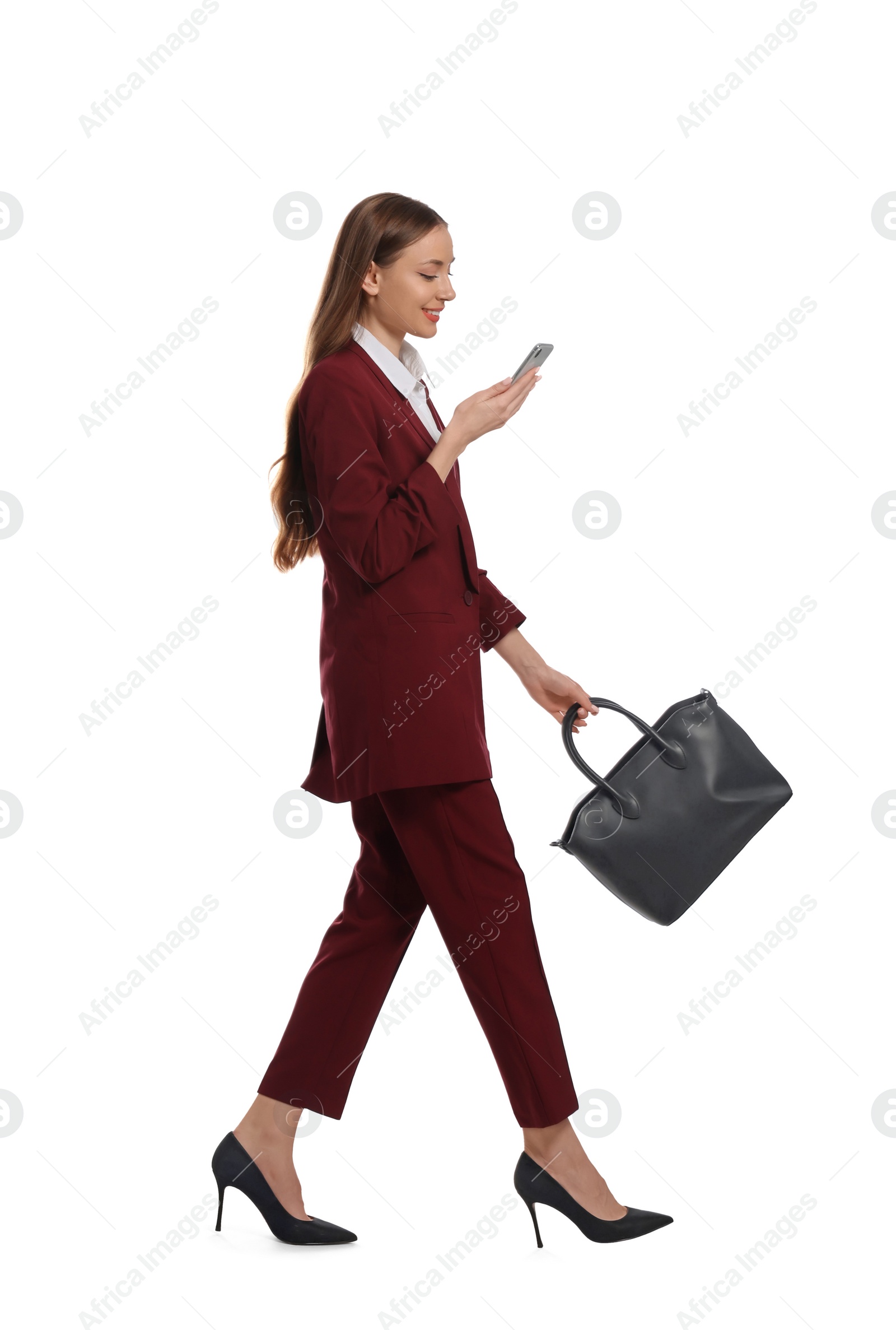 Photo of Young woman in burgundy suit using smartphone while walking on white background