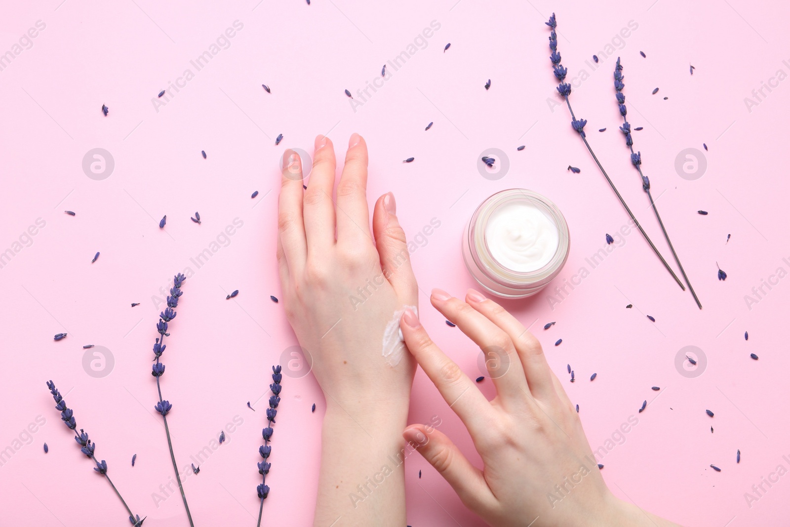 Photo of Woman applying hand cream and lavender flowers on pink background, top view