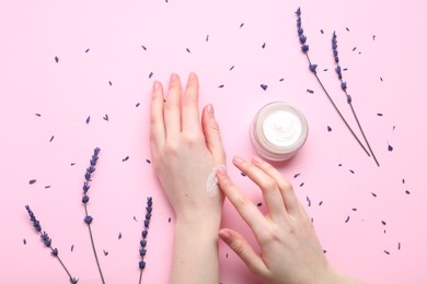 Woman applying hand cream and lavender flowers on pink background, top view