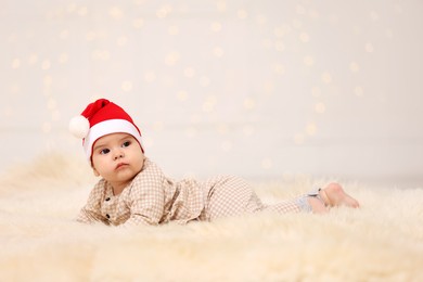 Cute baby in Santa hat on fluffy carpet against blurred festive lights, space for text. Winter holiday