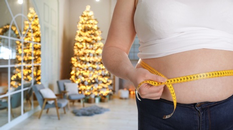 Image of Overweight woman measuring her waist in room with Christmas tree after holidays, closeup