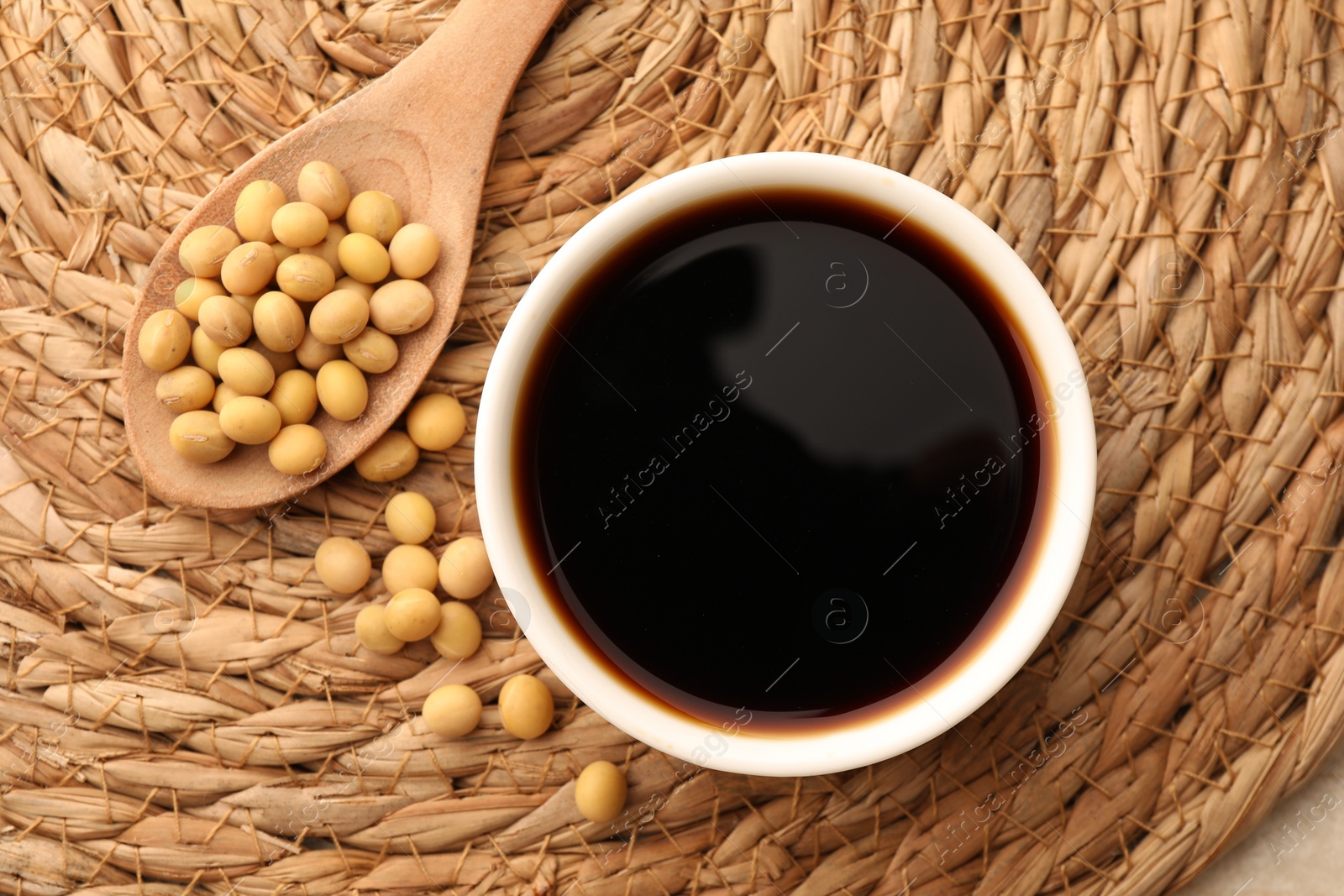 Photo of Soy sauce in bowl and soybeans on wicker mat, flat lay