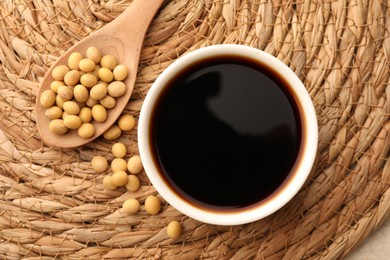 Photo of Soy sauce in bowl and soybeans on wicker mat, flat lay