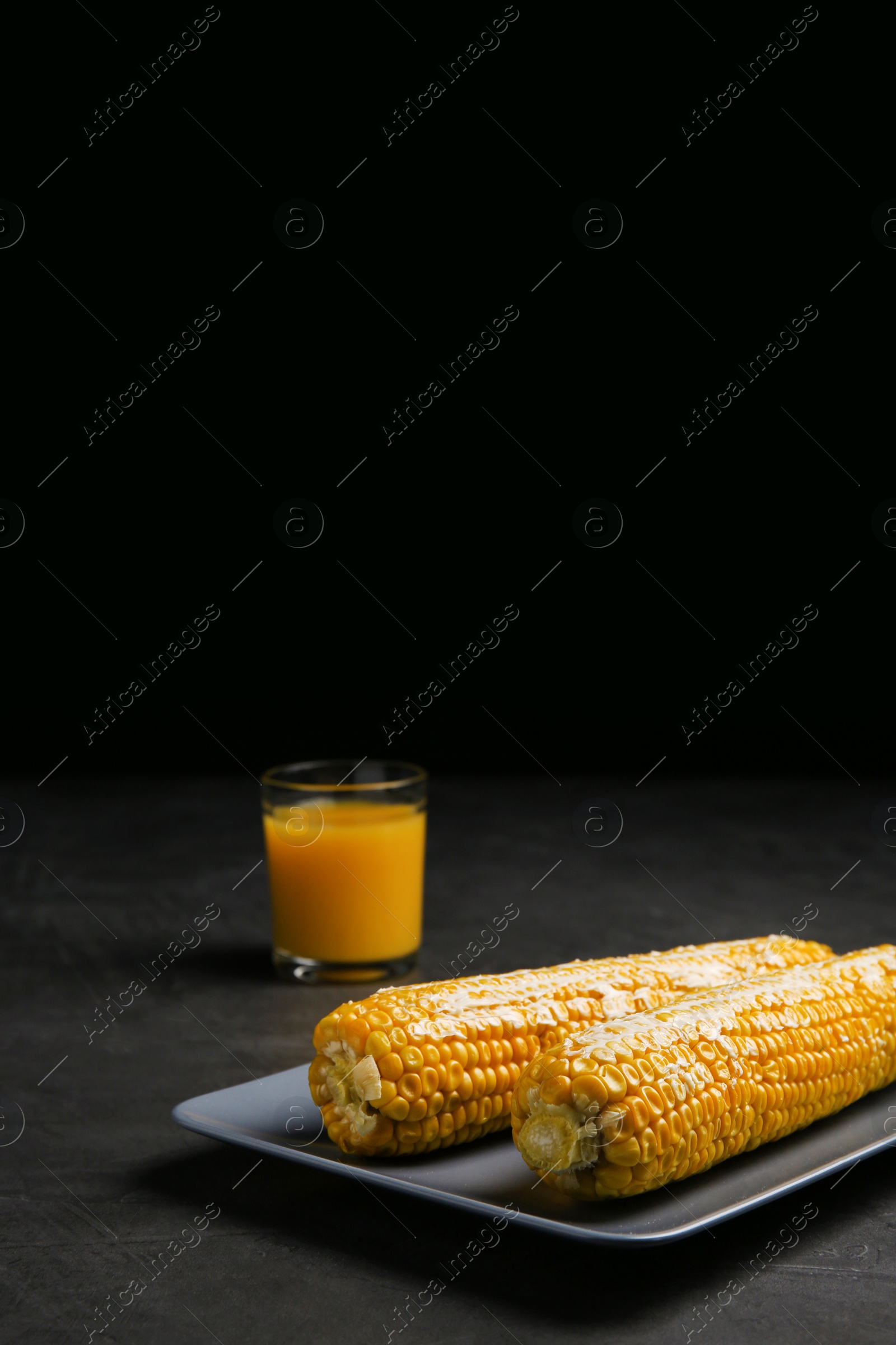 Photo of Delicious boiled corn cobs with butter on gray table against black background. Space for text