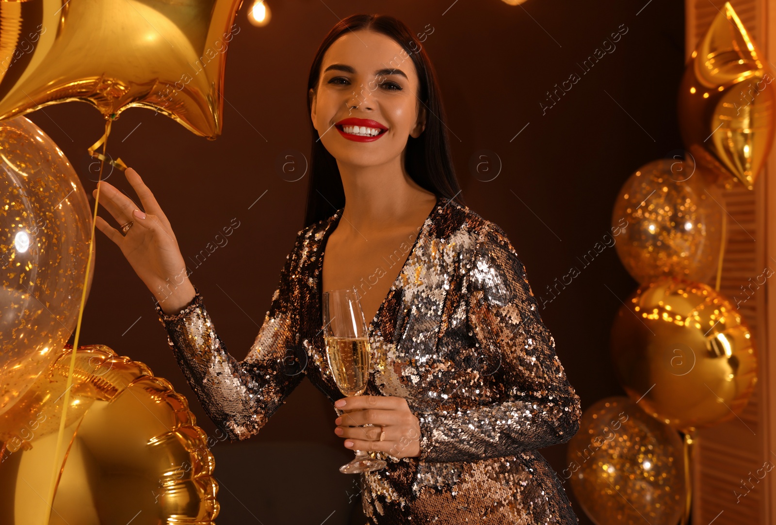 Photo of Beautiful young woman with glass of sparkling wine near golden balloons indoors. New Year celebration