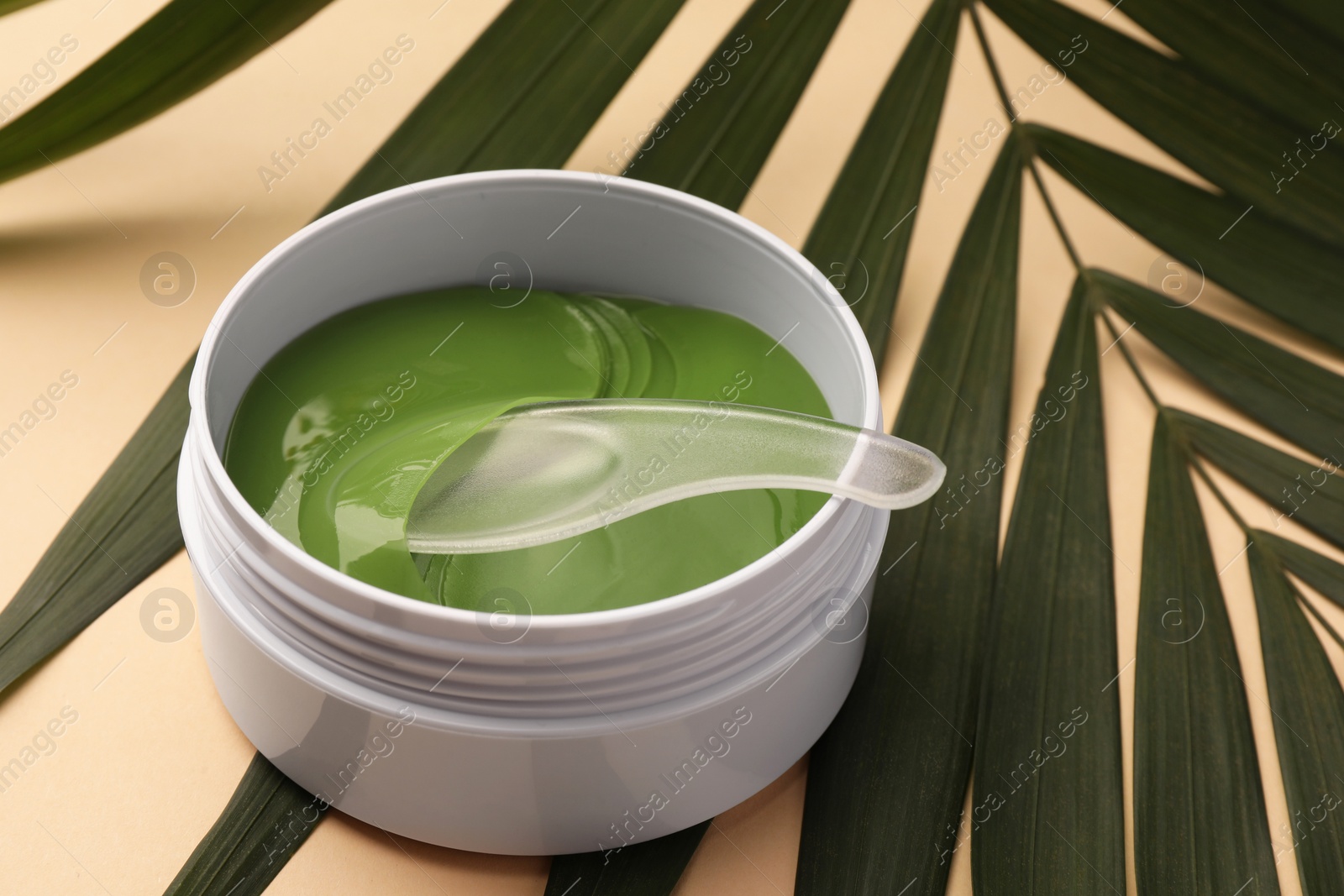 Photo of Jar of under eye patches with spoon and palm leaves on beige background, closeup. Cosmetic product