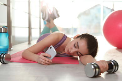 Lazy young woman with dumbbell and smartphone on yoga mat indoors