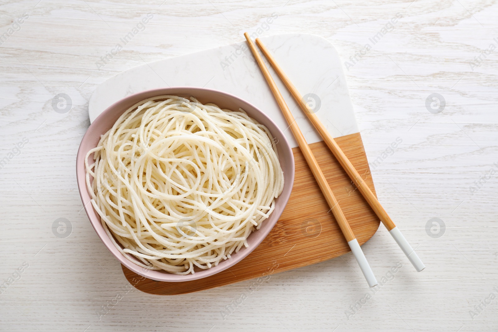 Photo of Bowl of tasty cooked rice noodles and chopsticks on white wooden table, top view