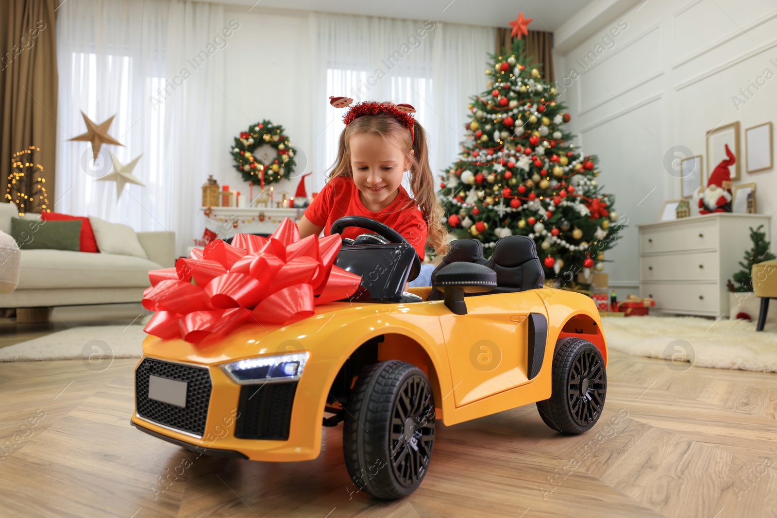 Photo of Cute little girl playing with toy car in room decorated for Christmas