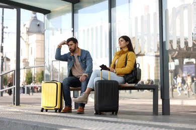 Photo of Being late. Worried couple with suitcases sitting at bus station outdoors