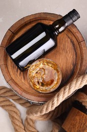 Whiskey with ice cubes in glass, bottle, wooden barrels and rope on grey table, top view