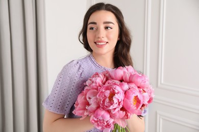 Photo of Beautiful young woman with bouquet of pink peonies indoors