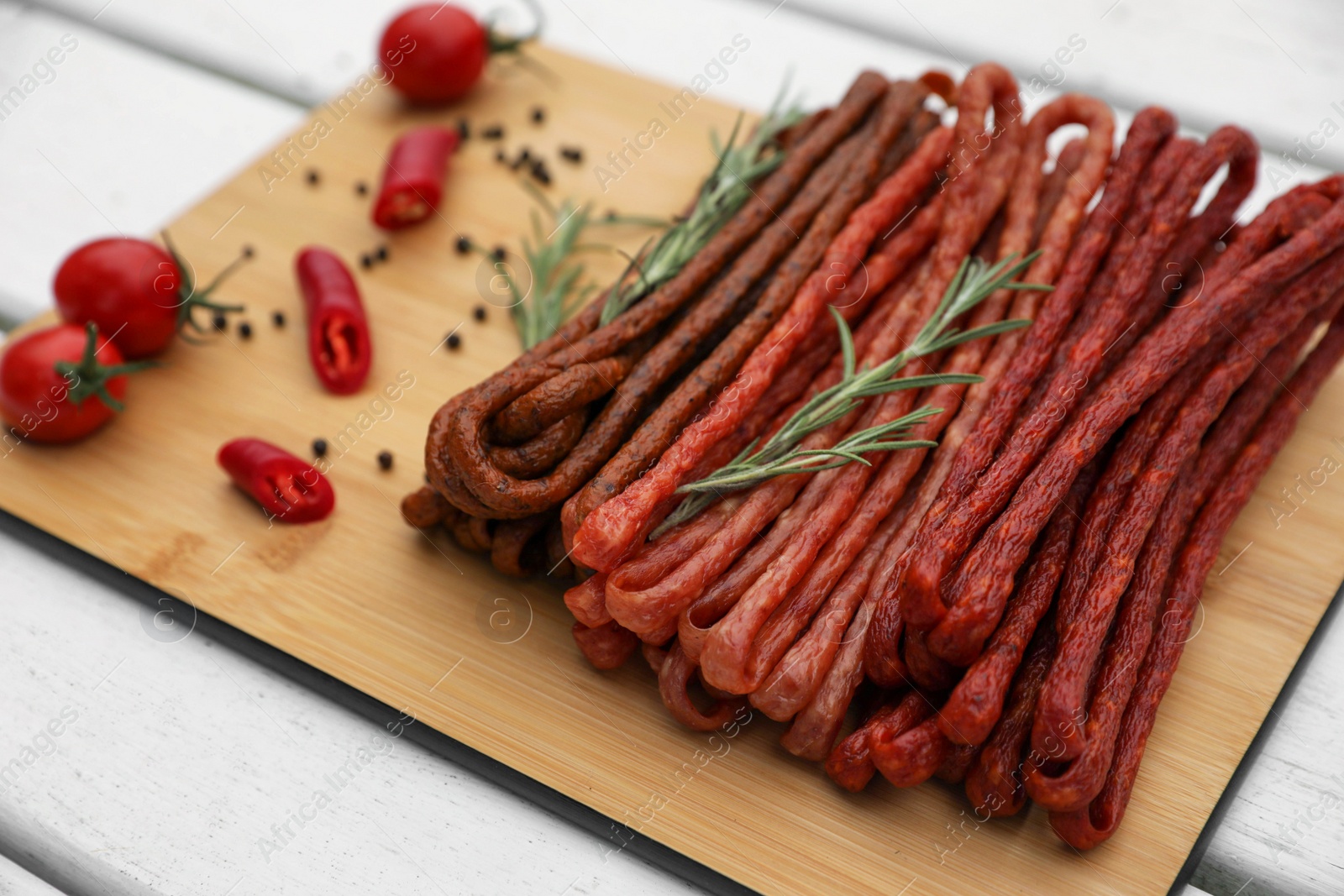 Photo of Delicious kabanosy with rosemary, peppercorn, chilli and tomatoes on white wooden table, closeup