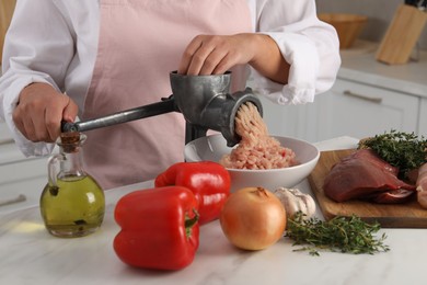 Woman making chicken mince with metal meat grinder at white table in kitchen, closeup