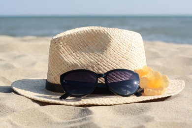 Photo of Hat with beautiful sunglasses and flowers on sand near sea, closeup