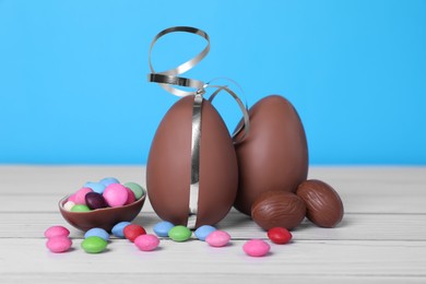 Photo of Delicious chocolate eggs and colorful candies on white wooden table against light blue background, closeup