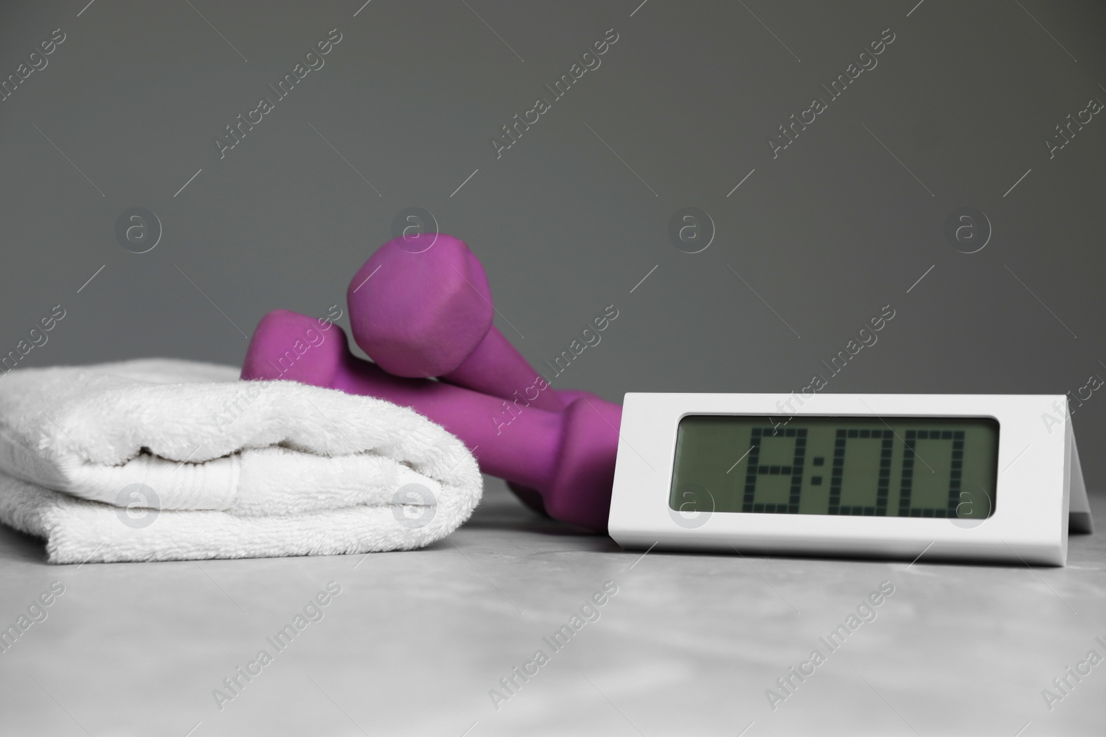 Photo of Alarm clock, dumbbells and towel on table against grey background. Morning exercise