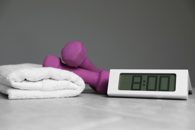 Alarm clock, dumbbells and towel on table against grey background. Morning exercise