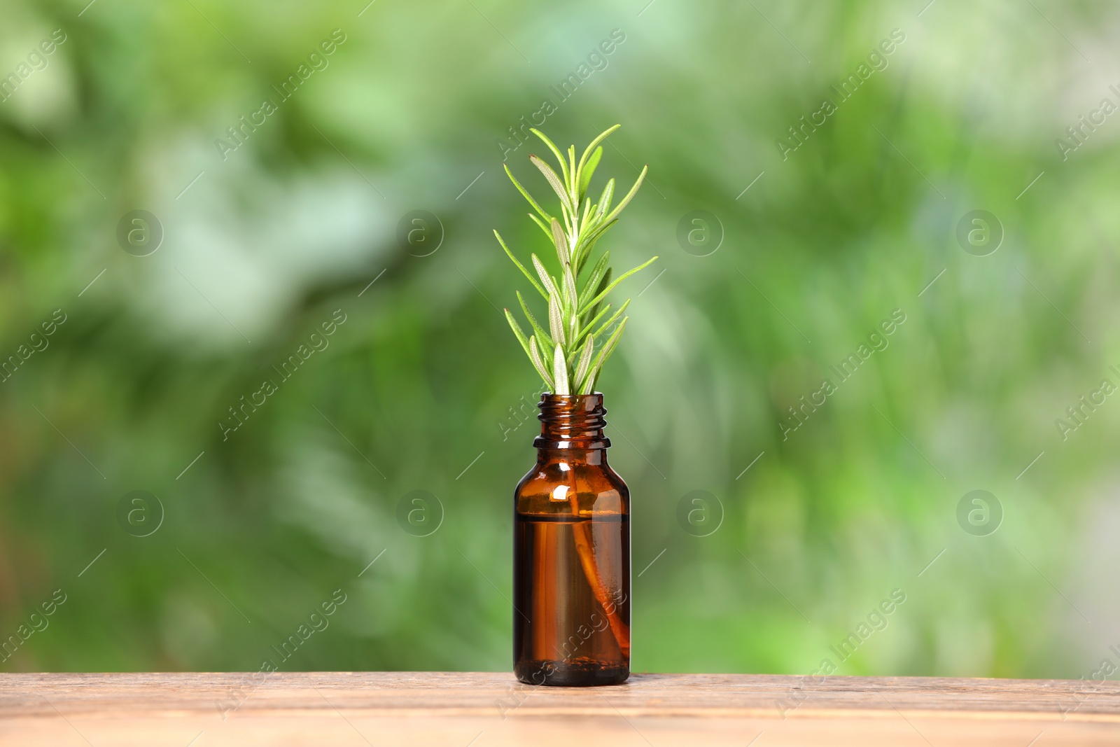 Photo of Bottle with essential oil and rosemary on wooden table against blurred green background