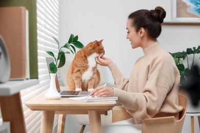 Photo of Woman writing in notebook and petting cute cat at home