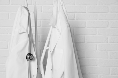 Photo of Medical uniforms and stethoscope hanging on rack near white brick wall. Space for text