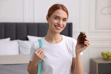 Photo of Happy young woman with bottle of pills and measuring tape in room. Weight loss