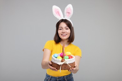 Photo of Easter celebration. Happy woman with bunny ears and wicker basket full of painted eggs on grey background