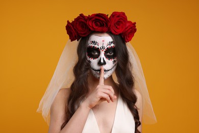 Photo of Young woman in scary bride costume with sugar skull makeup and flower crown showing shush gesture on orange background. Halloween celebration