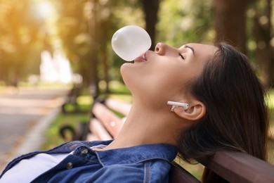 Photo of Beautiful young woman with wireless headphones blowing chewing gum on wooden bench outdoors