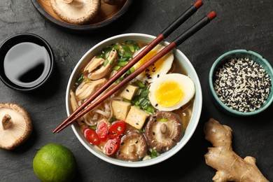 Photo of Bowl of delicious ramen, ingredients and chopsticks on black table, flat lay. Noodle soup