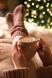 Photo of Woman holding cup of delicious hot drink near Christmas tree indoors, closeup