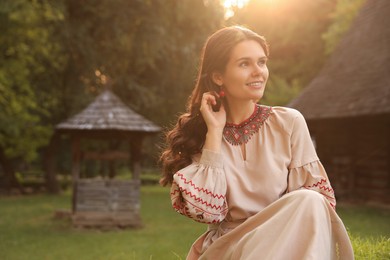 Photo of Beautiful woman wearing embroidered dress in village on sunny day, space for text. Ukrainian national clothes