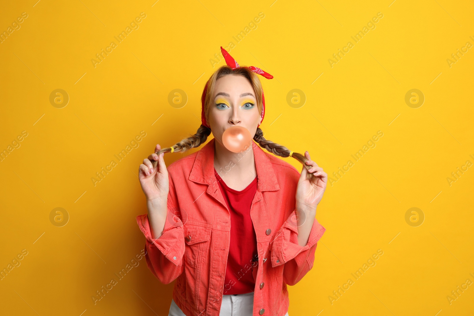 Photo of Fashionable young woman with braids and bright makeup blowing bubblegum on yellow background