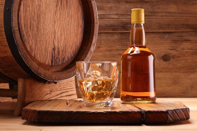 Whiskey with ice cubes in glass, bottle and barrel on wooden table