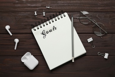 Photo of Notebook with goal list, earphones, glasses and stationery on wooden table, flat lay