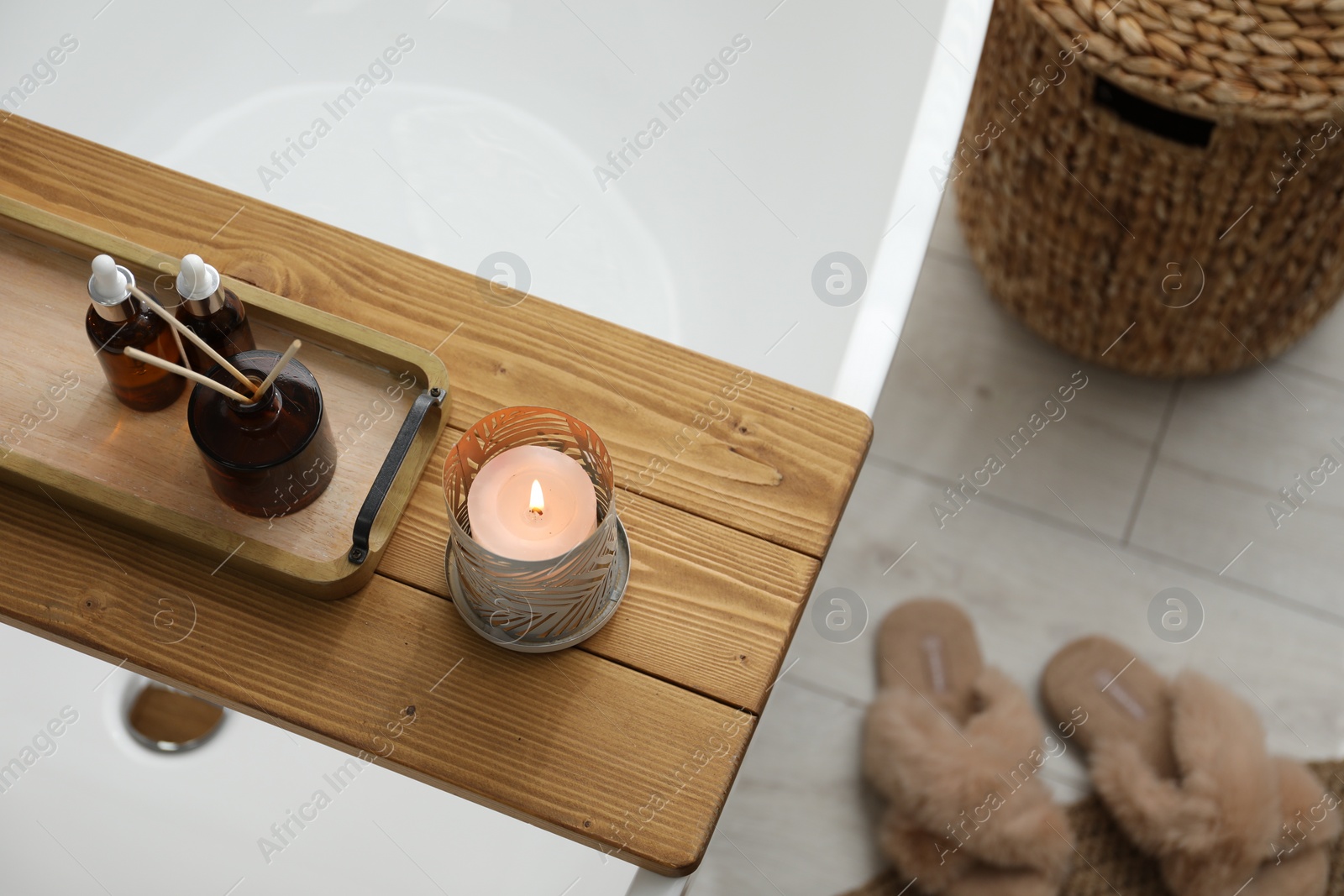 Photo of Wooden tray with cosmetic products, burning candle and reed air freshener on bath tub in bathroom, above view