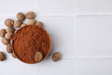 Photo of Nutmeg powder in bowl and seeds on white tiled table, flat lay. Space for text