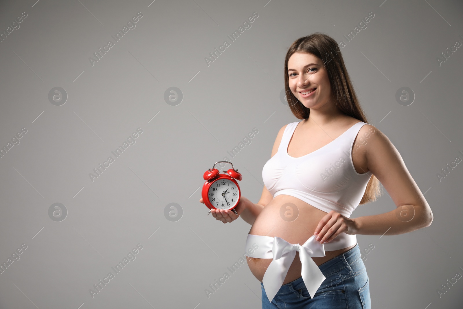 Photo of Young pregnant woman with alarm clock and bow on grey background, space for text. Time to give birth