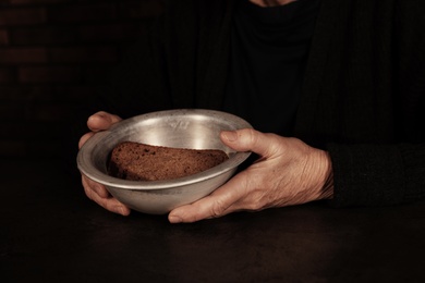 Photo of Poor mature woman holding bowl with bread at table, closeup