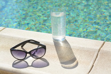 Photo of Stylish sunglasses and glass of water near outdoor swimming pool on sunny day