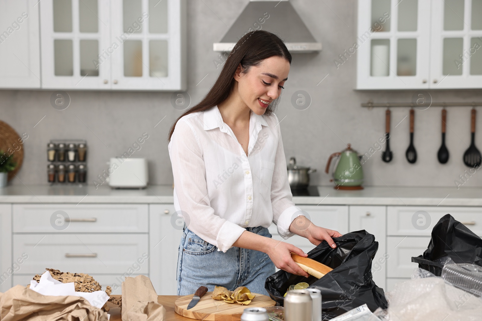 Photo of Garbage sorting. Woman putting food waste into plastic bag at table in kitchen