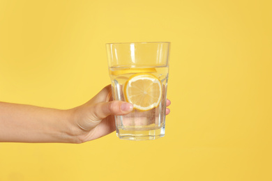 Photo of Young woman holding glass of lemon water on yellow background, closeup