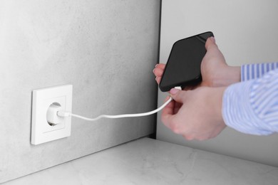 Woman plugging smartphone into power socket at white table indoors, closeup