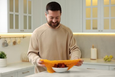 Man packing bowl of fresh grapes into beeswax food wrap at table in kitchen