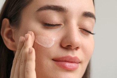 Young woman with dry skin applying cream onto her face on blurred background, closeup