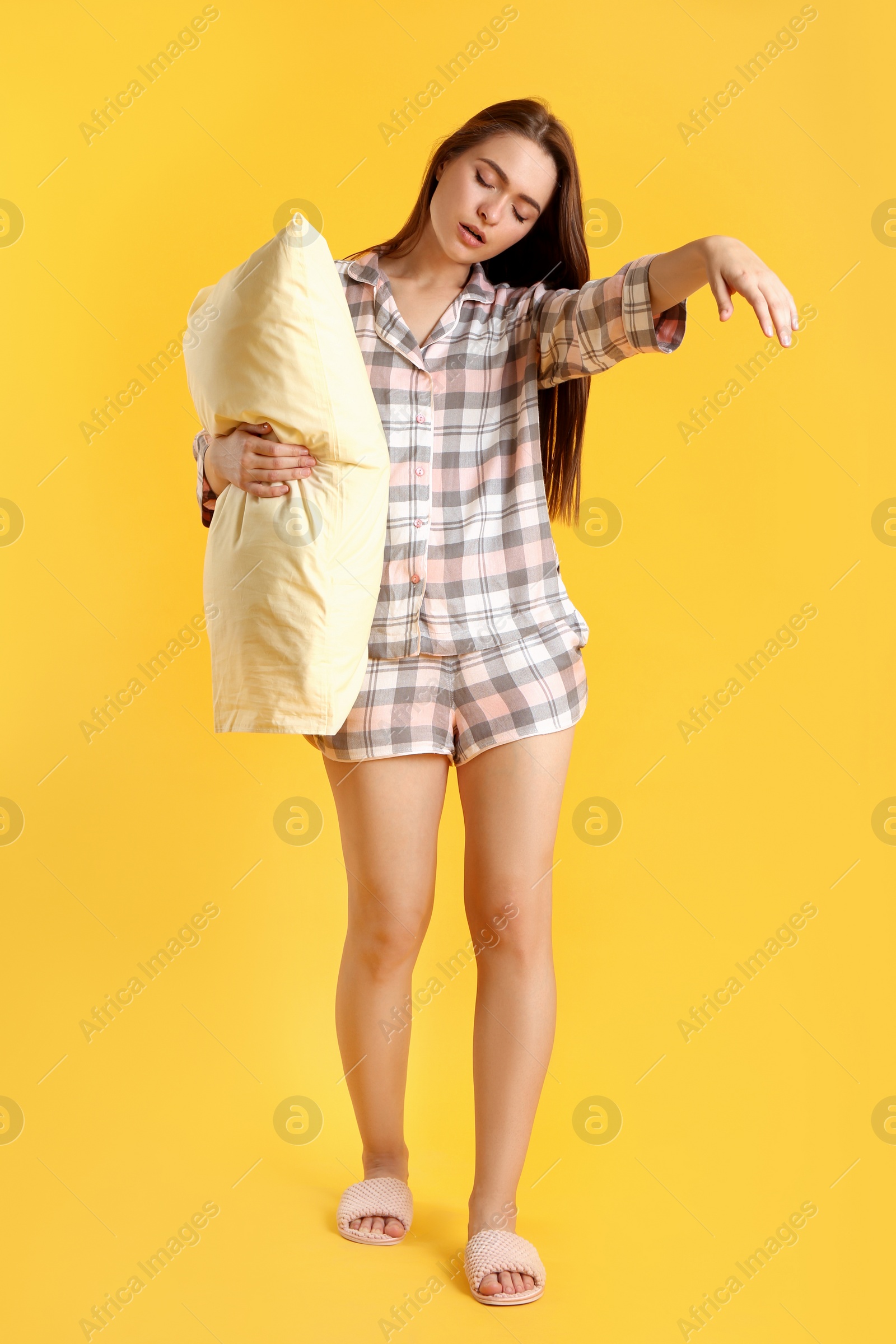 Photo of Young woman wearing pajamas and slippers with pillow in sleepwalking state on yellow background