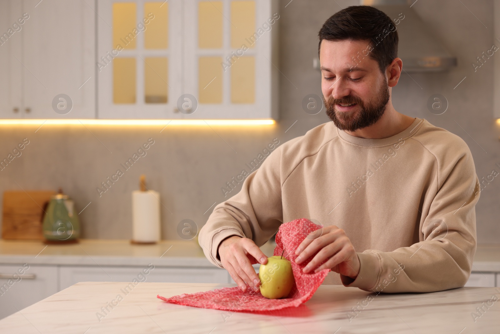 Photo of Man packing fresh apple into beeswax food wrap at table in kitchen. Space for text