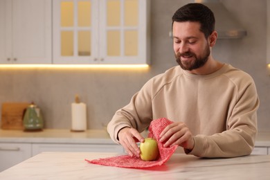 Photo of Man packing fresh apple into beeswax food wrap at table in kitchen. Space for text